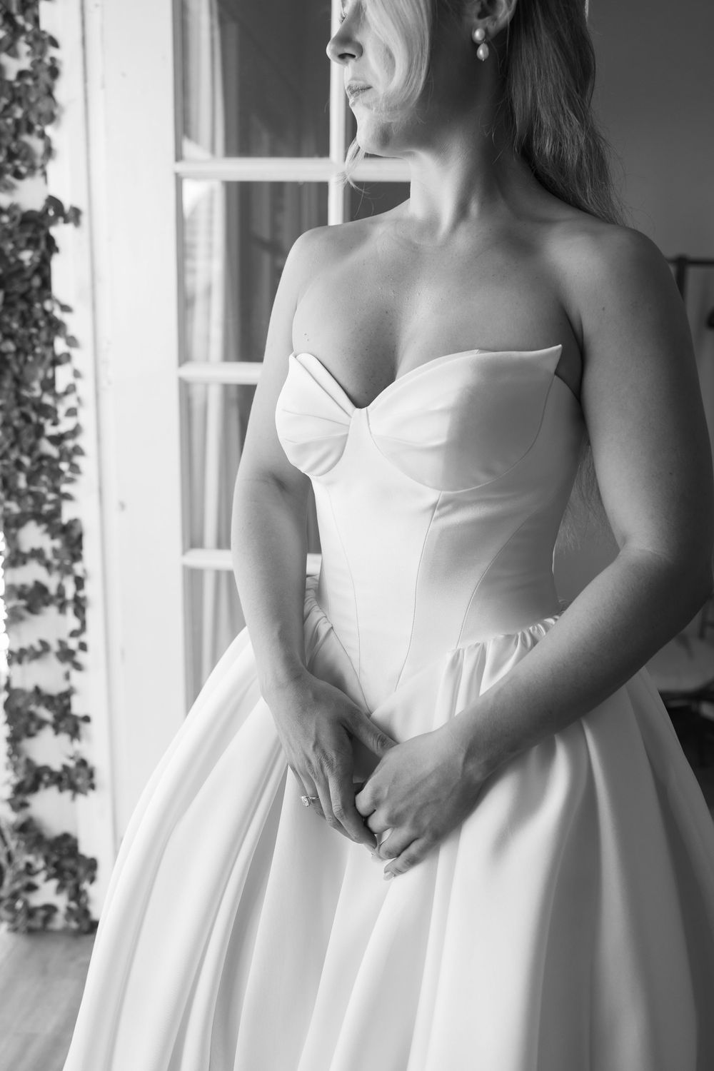Bride in strapless satin ball gown wedding dress standing by window in black and white photo.