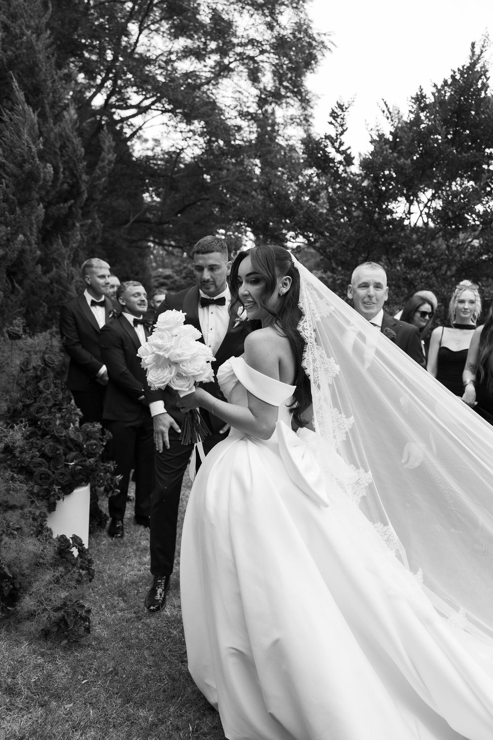 A joyful wedding party celebrates outdoors as the bride's white veil flows dramatically in the wind.