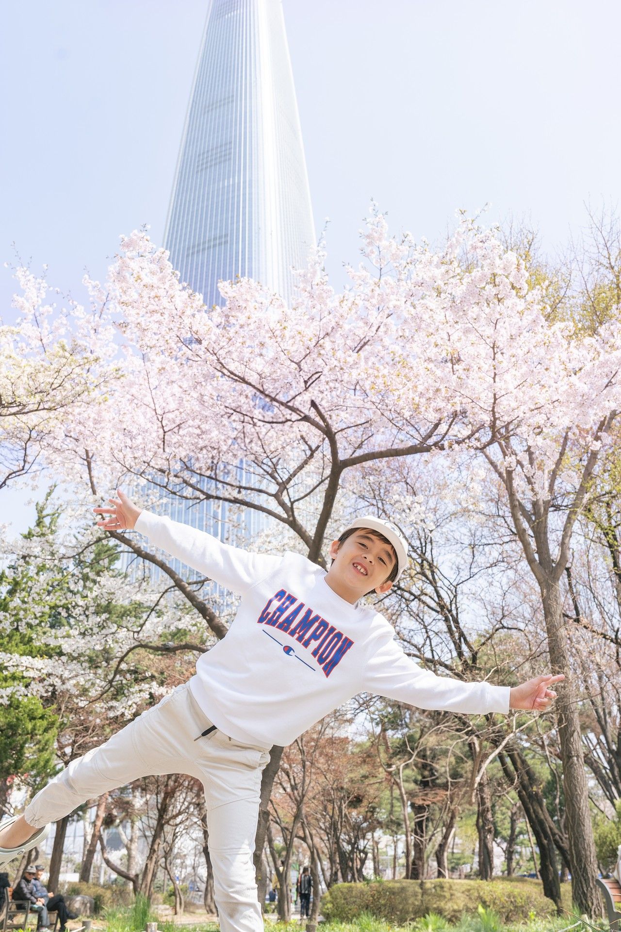 Cherry blossom trees bloom in a park with modern skyscrapers in the background as someone poses playfully.