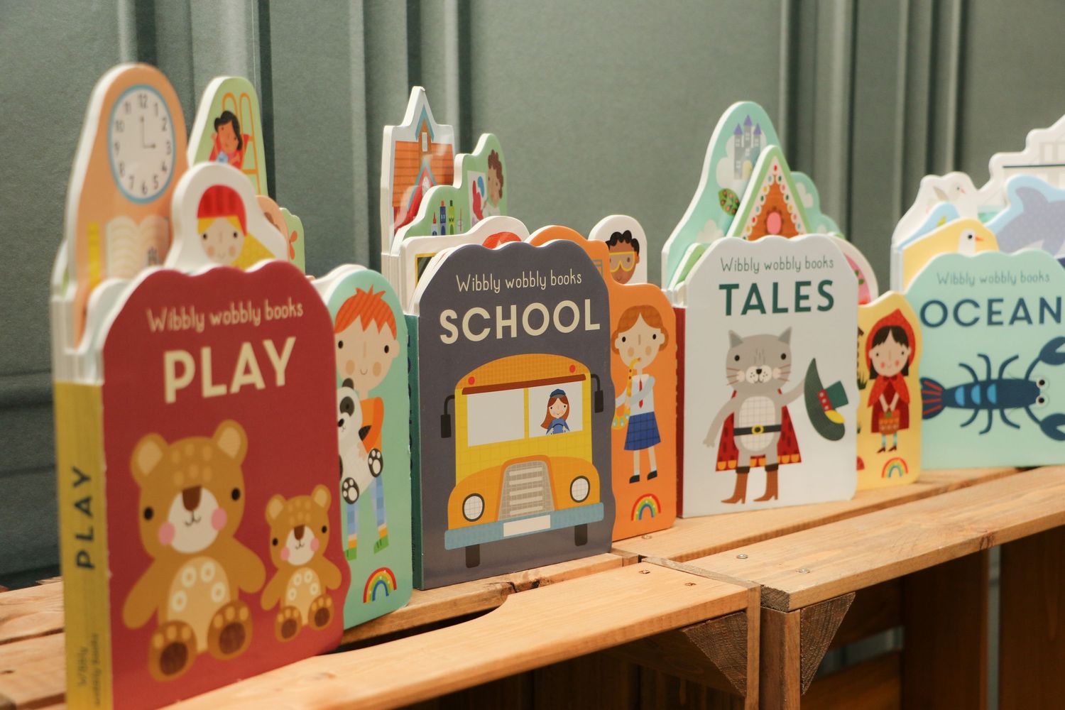 Colorful children's board books arranged in a row on a wooden shelf display educational titles about school, play and ocean themes.