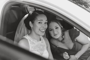 Two people in bridal attire share a joyful moment together inside a car, captured in black and white photography.