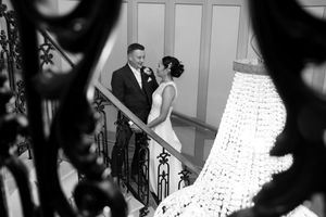 A black and white wedding photo captured on an elegant staircase with ornate iron railings and dramatic lighting.