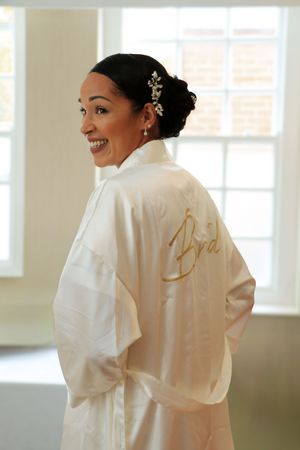 Woman in white silk robe with embroidered details getting ready in bright sunlit room.