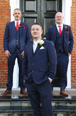A group of groomsmen in matching navy suits and red ties pose on stone steps in front of a brick building.
