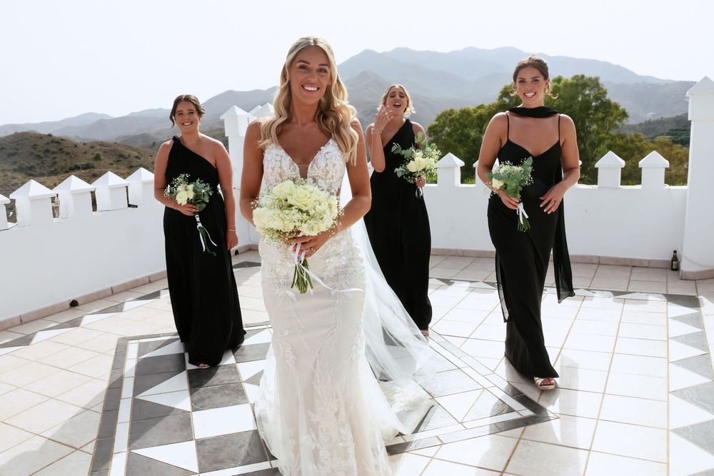Bride in lace gown holds white bouquet with bridesmaids in black dresses on rooftop with mountain views.