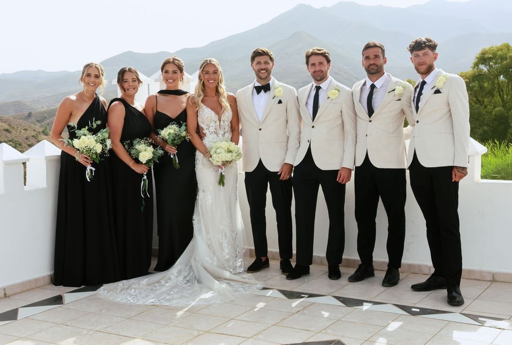 Wedding party posing outdoors with bride in white gown, bridesmaids in black dresses, and groomsmen in white jackets.
