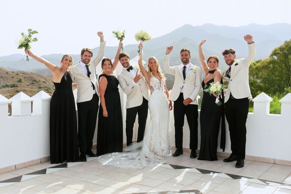 Wedding party in black and white attire celebrate with raised arms on a rooftop with scenic mountains.