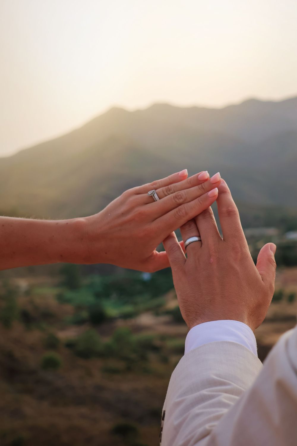 Couple touching hands with wedding rings at sunset, mountains in background.