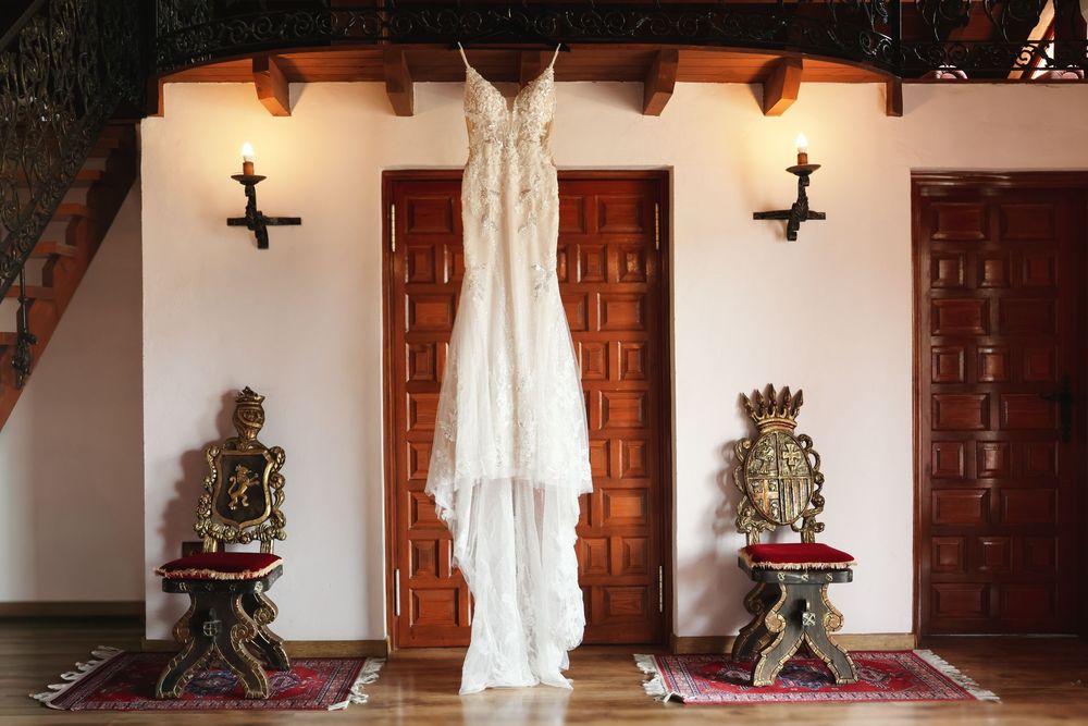Elegant lace wedding dress hanging between ornate golden chairs and wooden doors in a Spanish colonial interior.