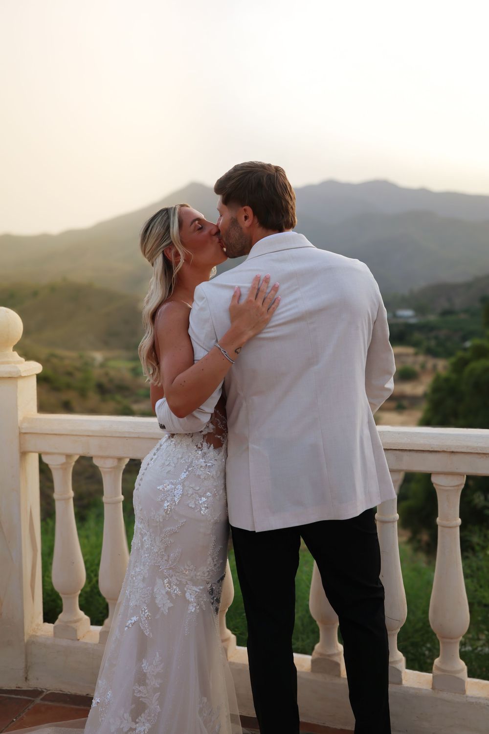 A bride and groom share a romantic kiss on a white balcony with scenic mountain views at sunset.