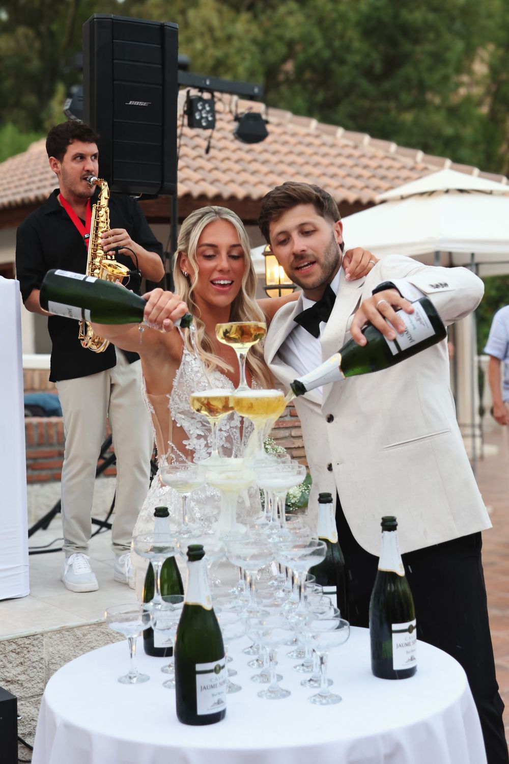 Bride and groom pouring champagne tower at wedding reception with saxophonist performing in background.