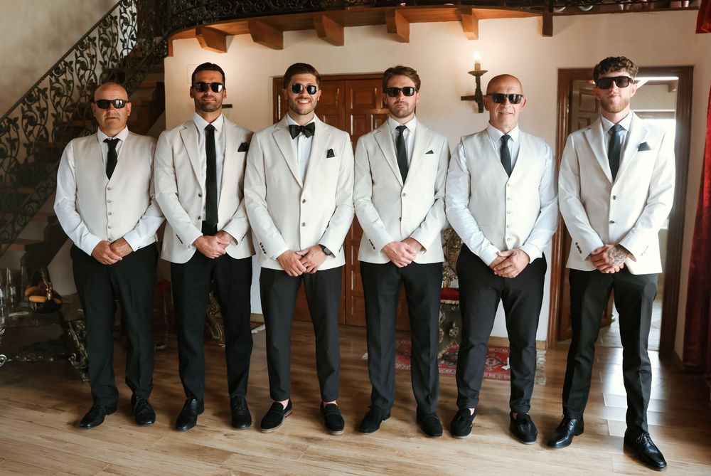 Six groomsmen in matching white tuxedo jackets, black ties, and sunglasses posing together at a wedding venue.