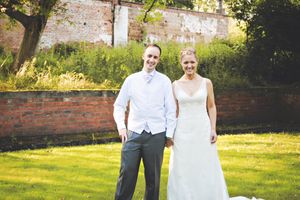 A wedding couple poses together in a sunny garden setting with brick walls and lush greenery in the background.