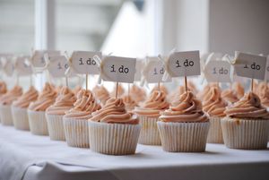 Wedding cupcakes with peach frosting and small 'I do' flags arranged in rows on a white tablecloth.