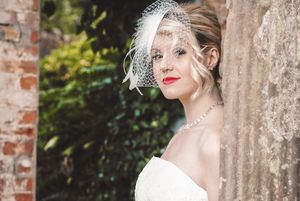 Bride in vintage-inspired lace gown poses elegantly against rustic brick wall in garden setting.