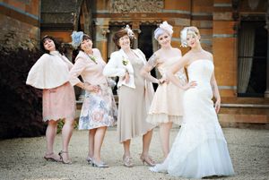 Group of bridesmaids in soft pink dresses and white gowns gather outside a brick building for wedding photos.