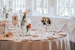 Elegant wedding reception tables decorated with white linens, floral centerpieces, and crystal glassware in a bright venue.