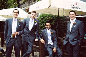 A group of groomsmen in black suits gather outdoors under white umbrellas and tents at a wedding celebration.
