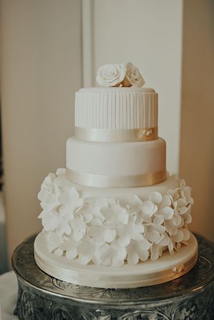 Three elegant white wedding cakes decorated with delicate floral details displayed on ornate silver platters.