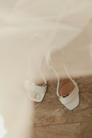 Close-up view of white leather bridal shoes with delicate straps and buttons on a beige carpet.