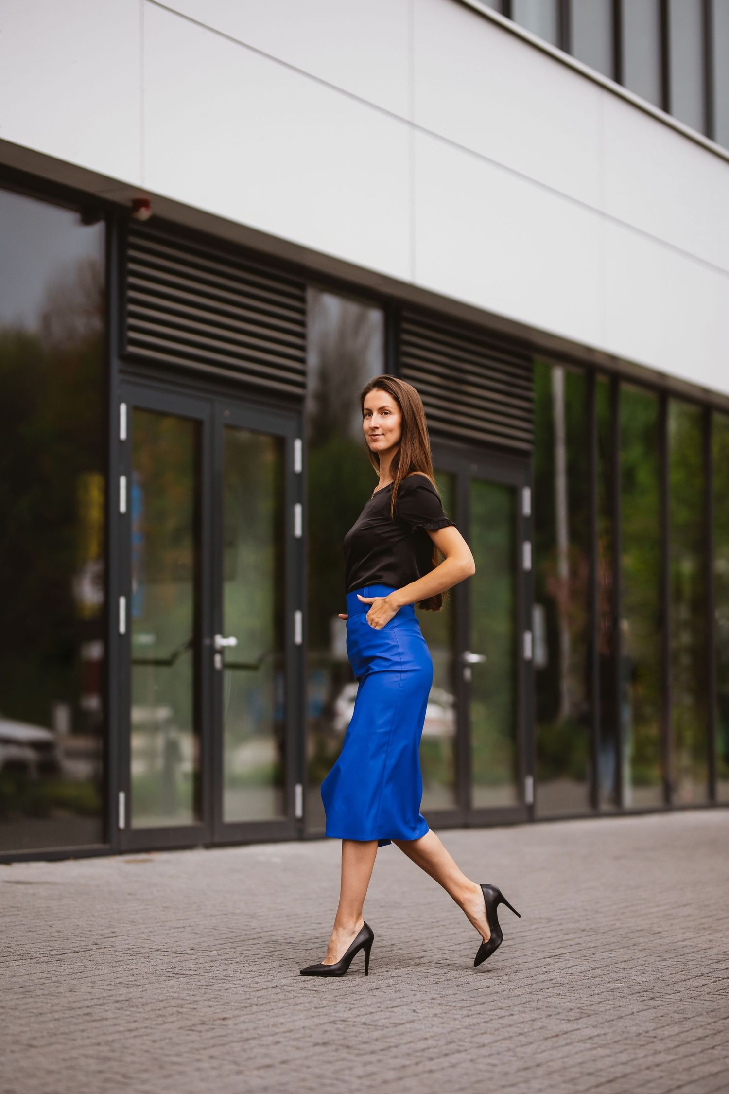 Elegantná žena v modrej sukni a čiernom tope kráča pred modernou budovou. Štýlové biznis fotenie v mestskom prostredí, Žilina.