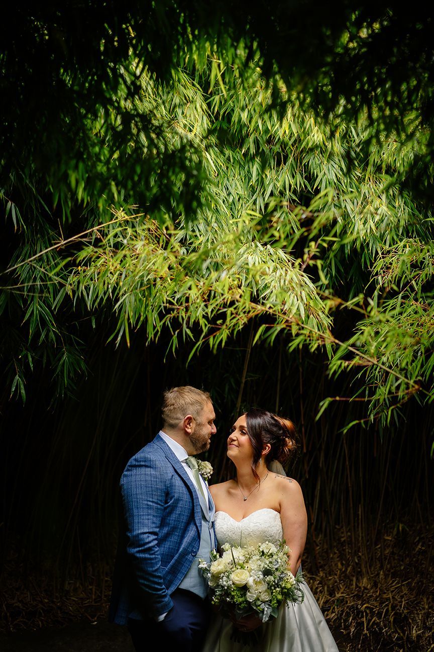 A couple shares a joyful moment under green bamboo leaves while holding a white floral bouquet.