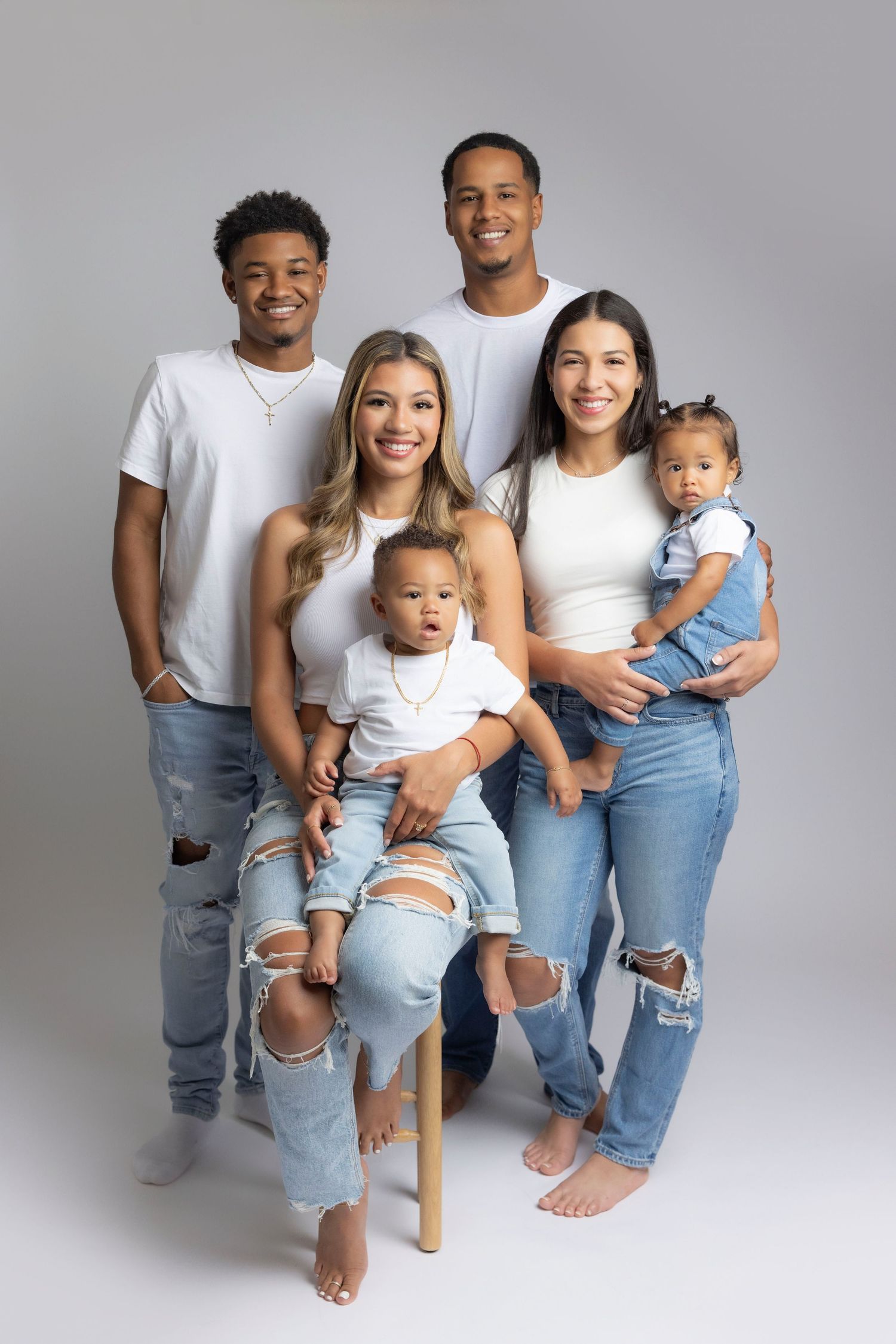 A family wearing matching white tops and ripped jeans poses together against a gray studio backdrop for portraits.