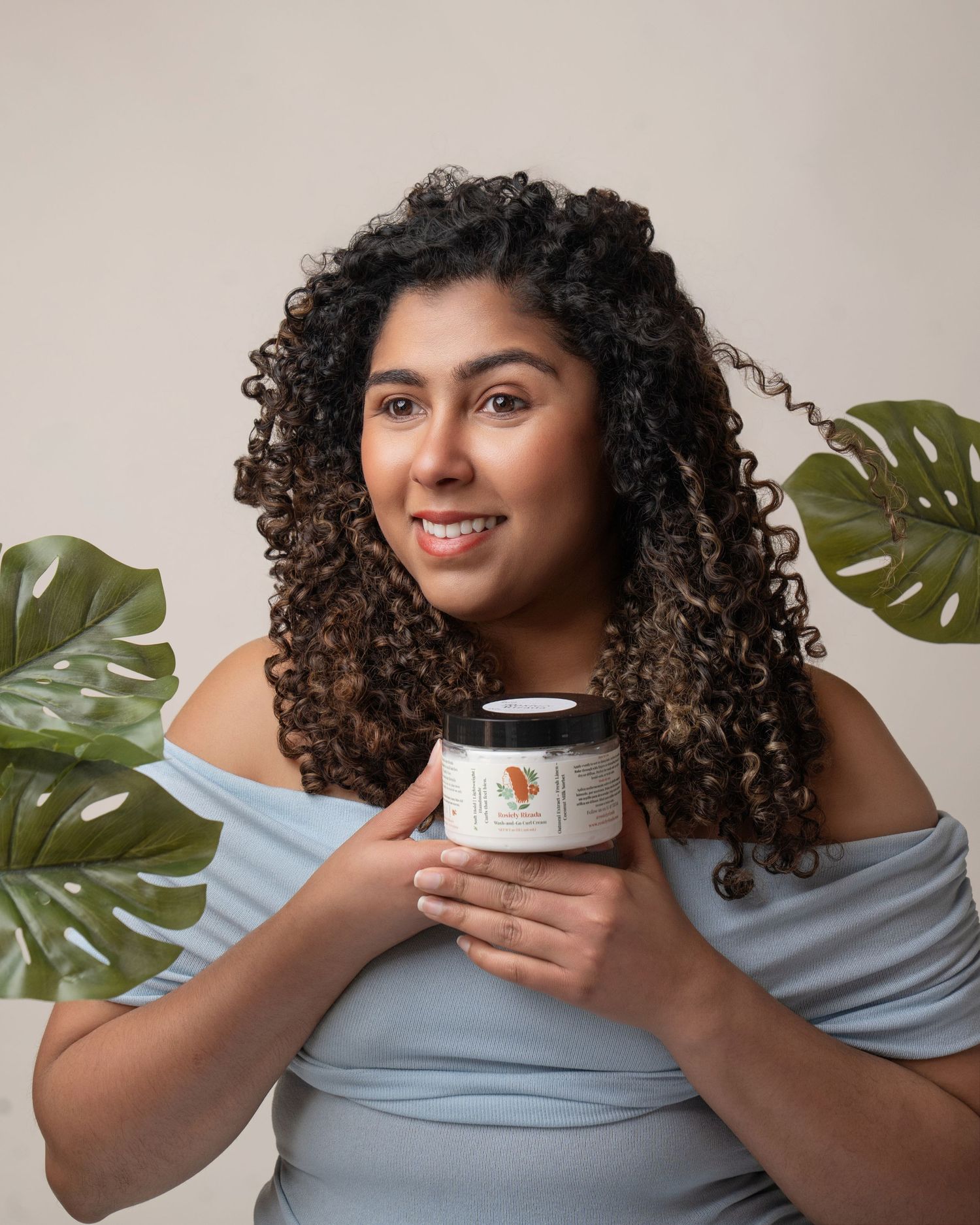 Person with curly hair holds a white skincare product jar against a light background with a monstera leaf.