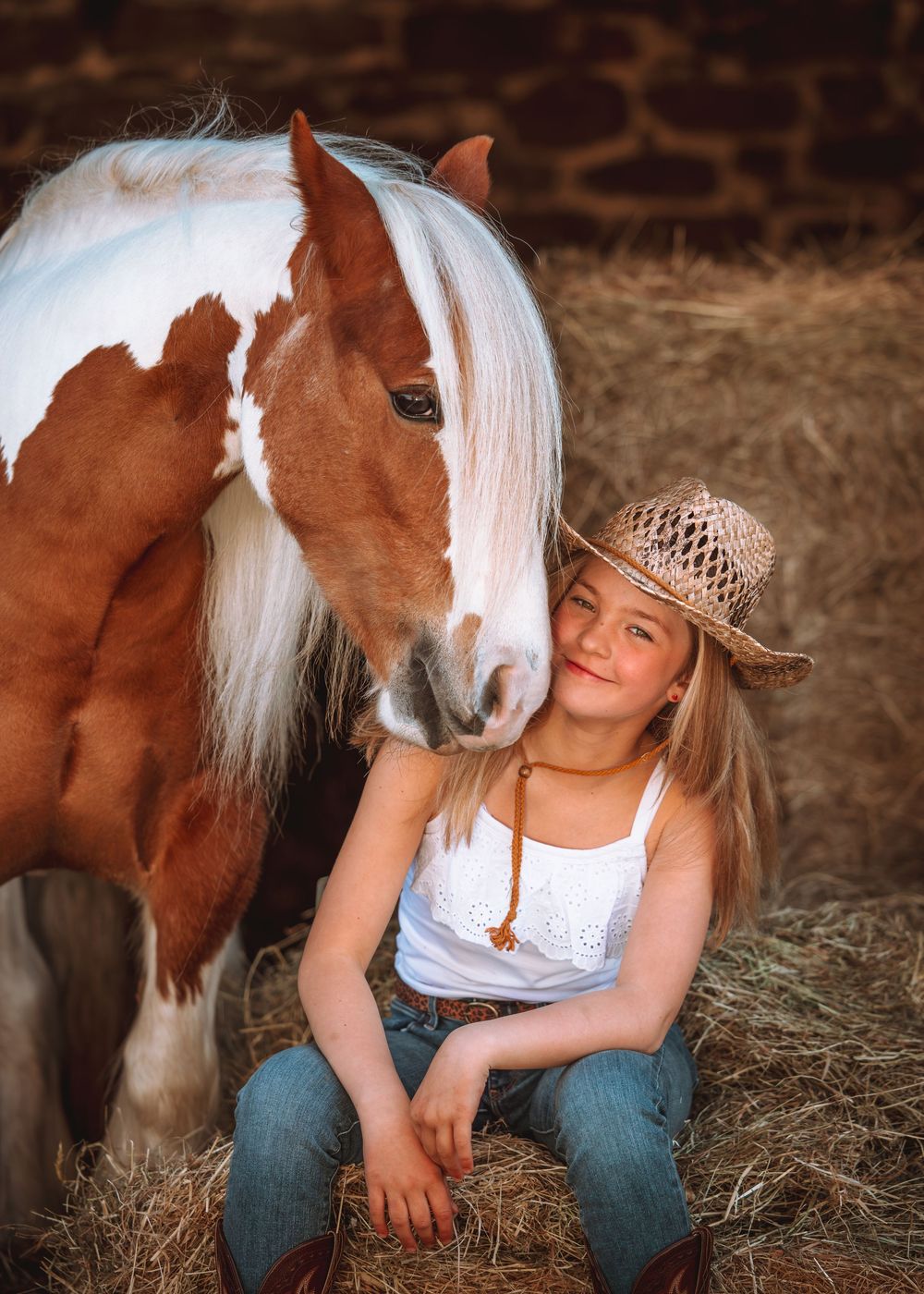 A brown and white pony with long mane stands in a rustic barn setting with hay on the ground.