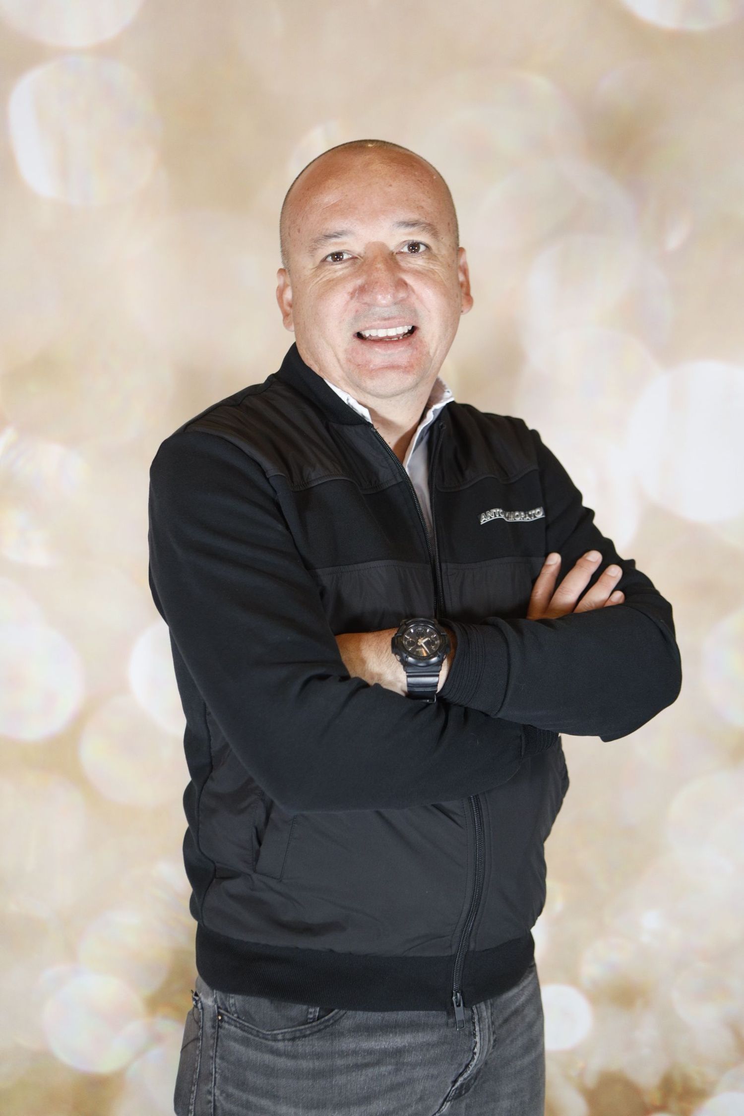 Professional portrait of someone in a black zip-up jacket with arms crossed against a light bokeh background.