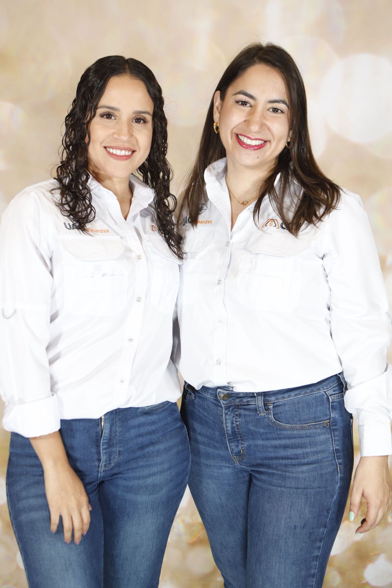 Two women wearing matching white button-down shirts and blue jeans pose together against a beige background.