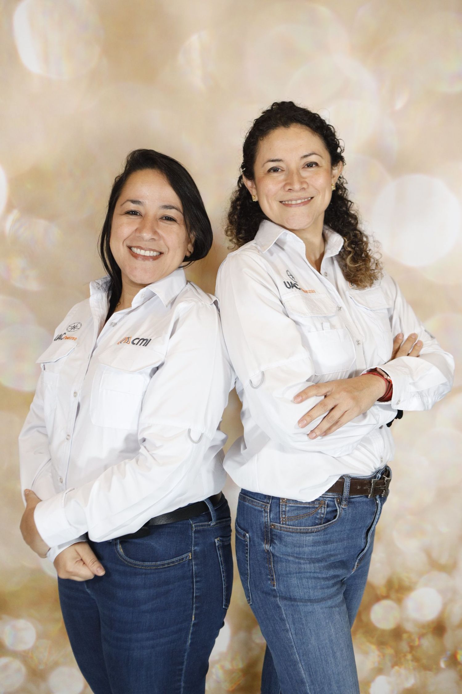 Two colleagues in matching white button-down shirts and jeans pose together against a sparkly beige backdrop.