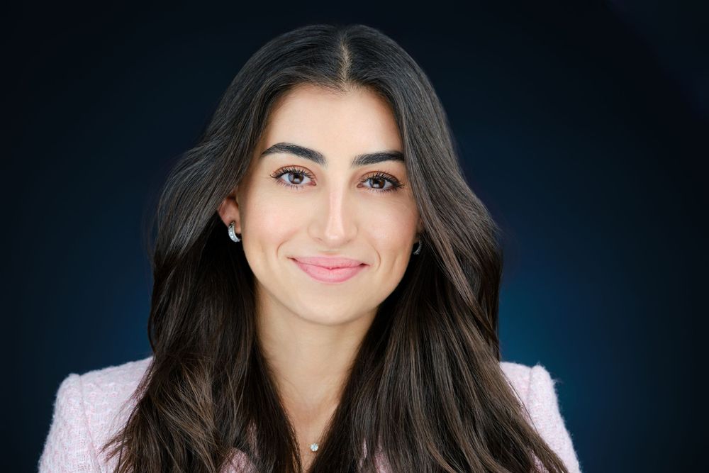 Professional headshot of a young woman with long dark hair wearing a pink blazer and delicate jewelry.
