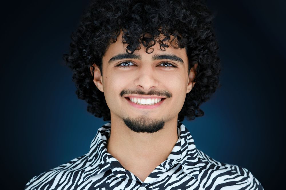 Smiling young man with curly dark hair and a beard wearing a zebra print shirt.