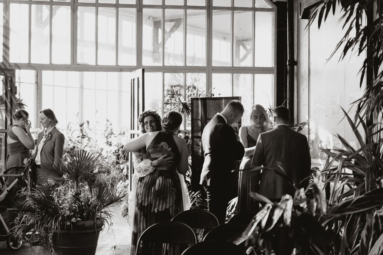 A black and white panoramic photograph shows people gathered in a greenhouse or conservatory with large windows.