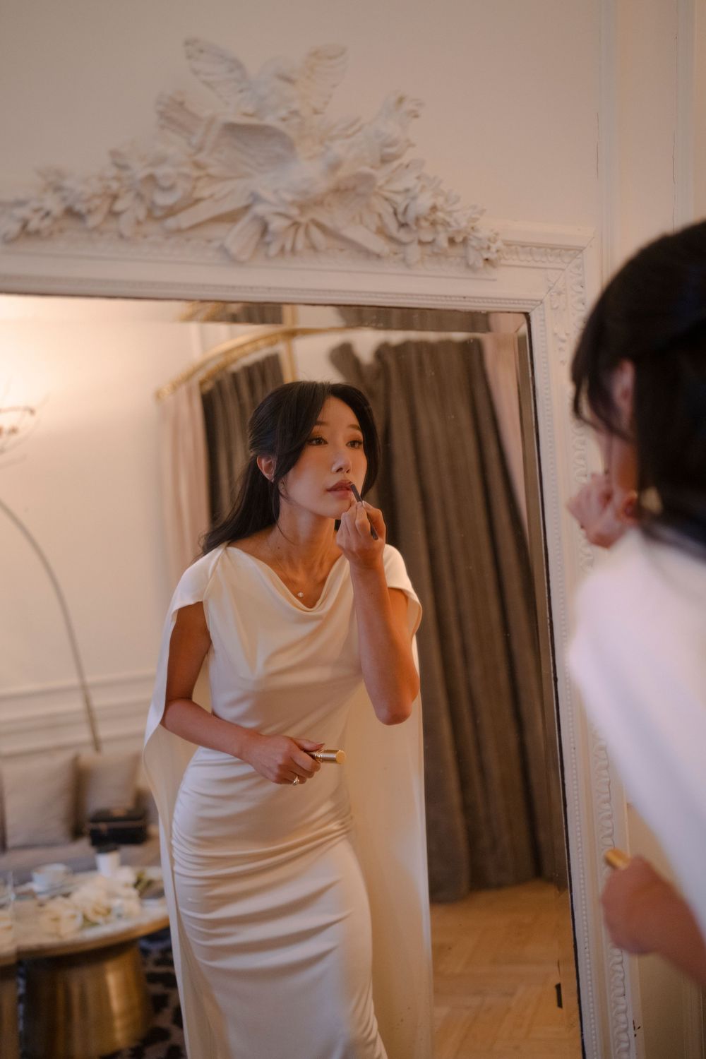 A person in a white dress spins and dances joyfully in front of a mirror in an elegant room.