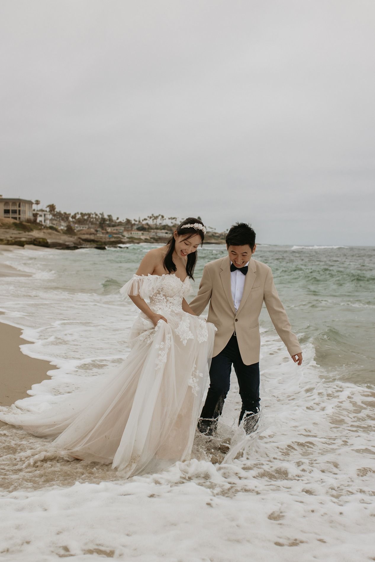 Couple in wedding attire playfully splashing through ocean waves at beach during romantic photoshoot at Windansea Beach in La Jolla.