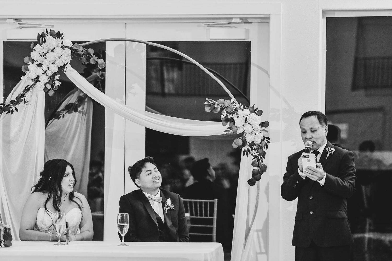 the bride and groom laugh while the groom's father toasts them