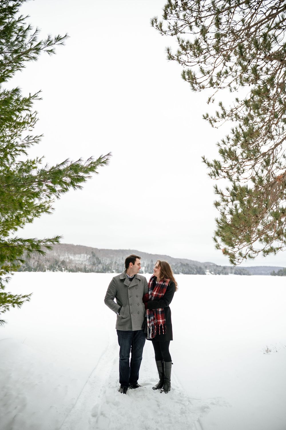 A couple enjoys a romantic winter moment in the snow with evergreen trees in the background.