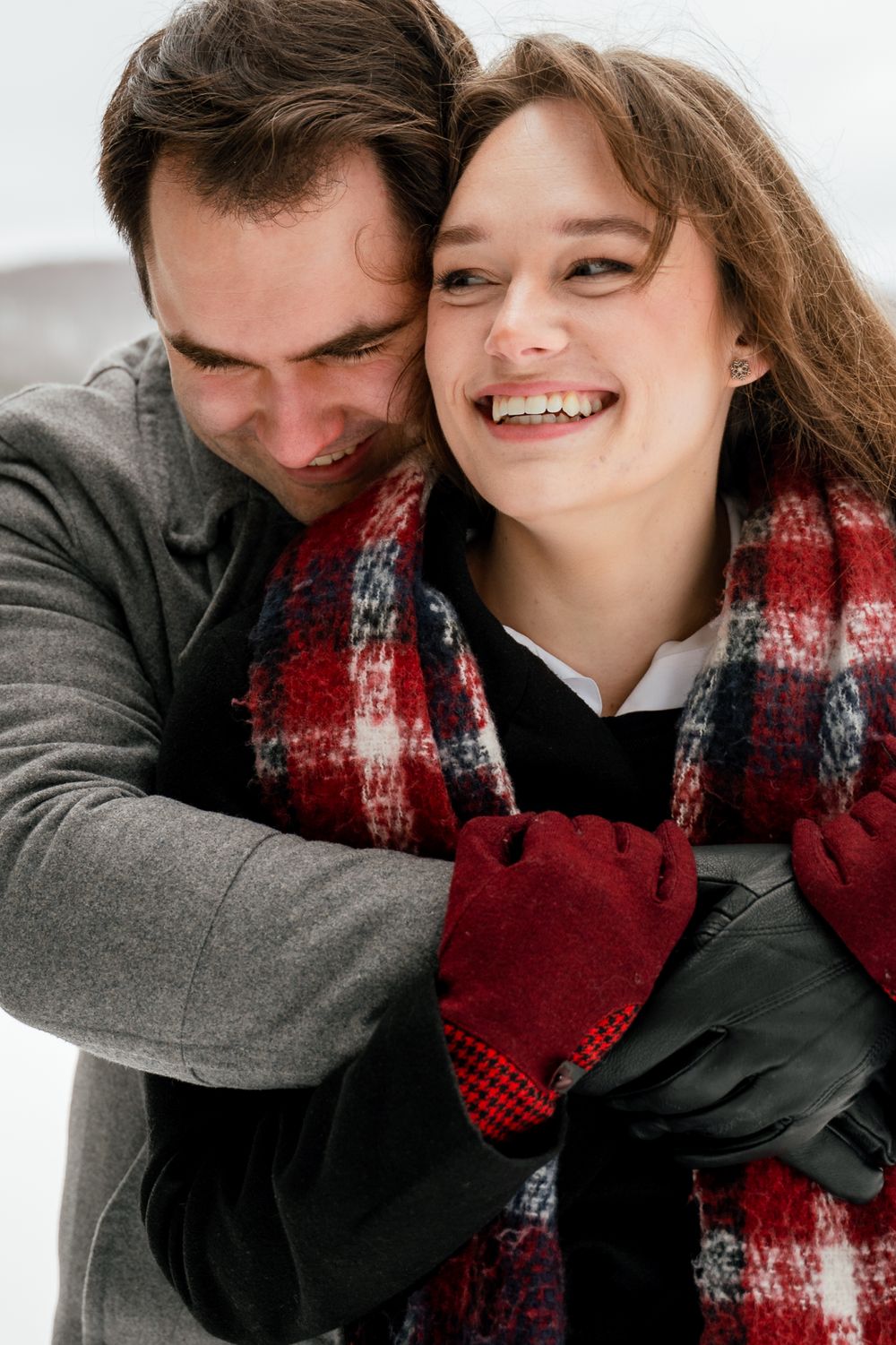 A couple wearing winter attire share an intimate moment while wrapped in a red plaid scarf.