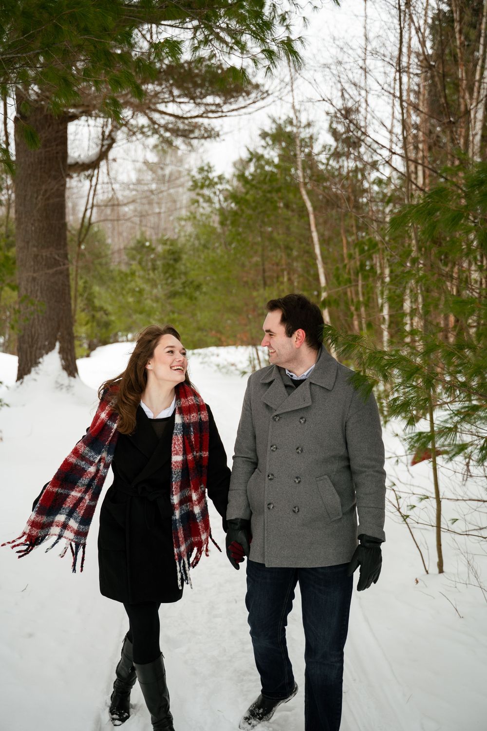 A couple walks together through a snowy forest wearing winter coats and a plaid scarf.