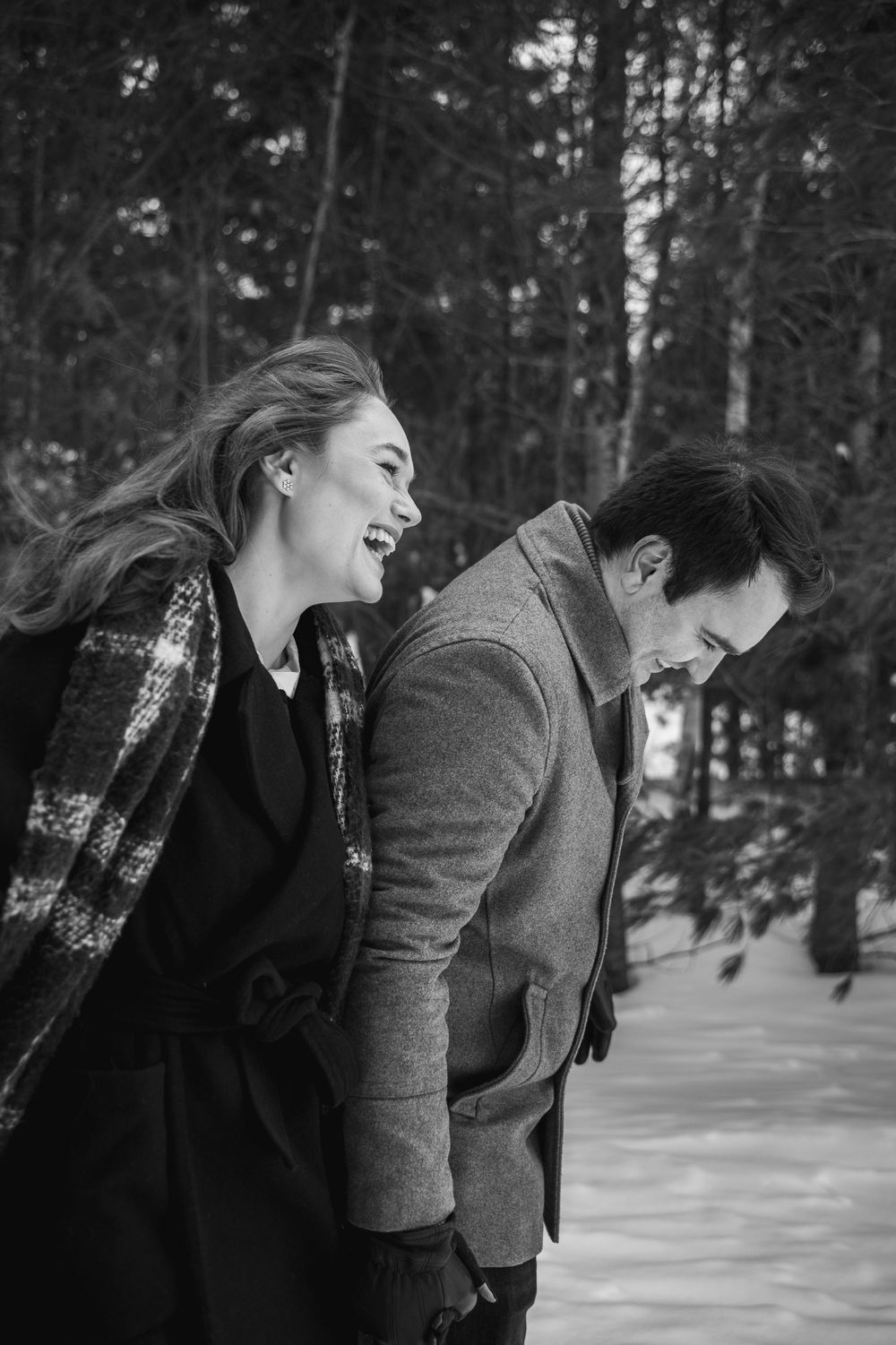 Two people share a joyful moment laughing together in a snowy winter forest setting.