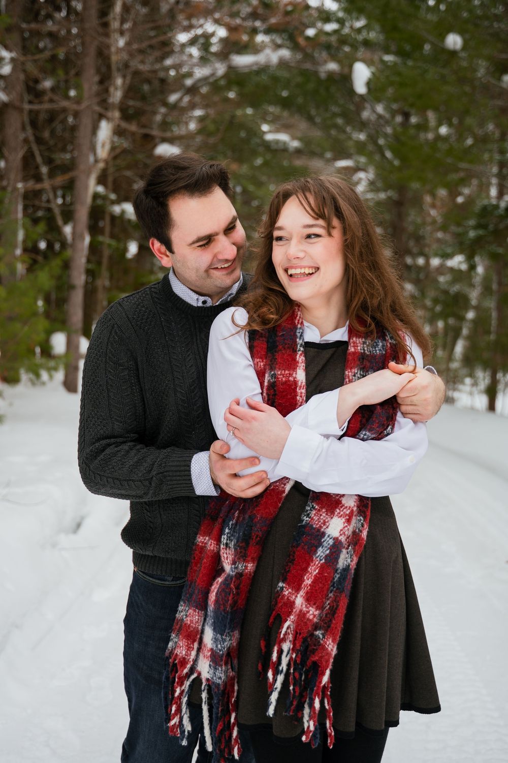 A playful winter engagement photoshoot in a snowy forest with a couple wearing scarves.