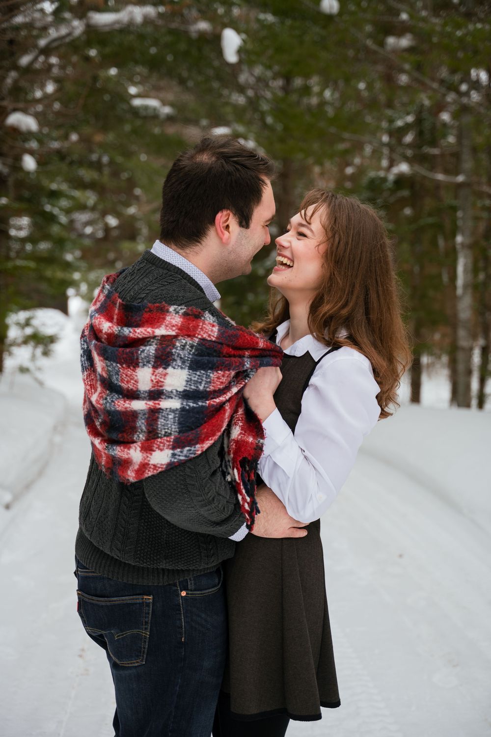 Playful winter couple photos with a red plaid scarf against a snowy forest backdrop.