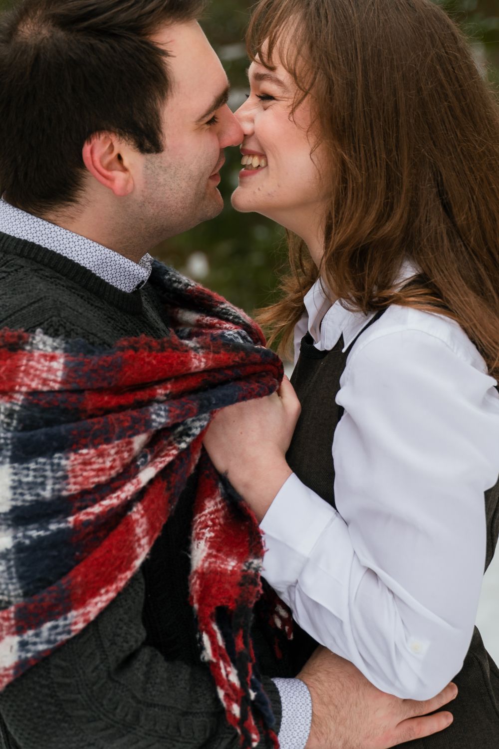 A couple shares a romantic moment wrapped in a red and navy plaid blanket during an outdoor photo session.