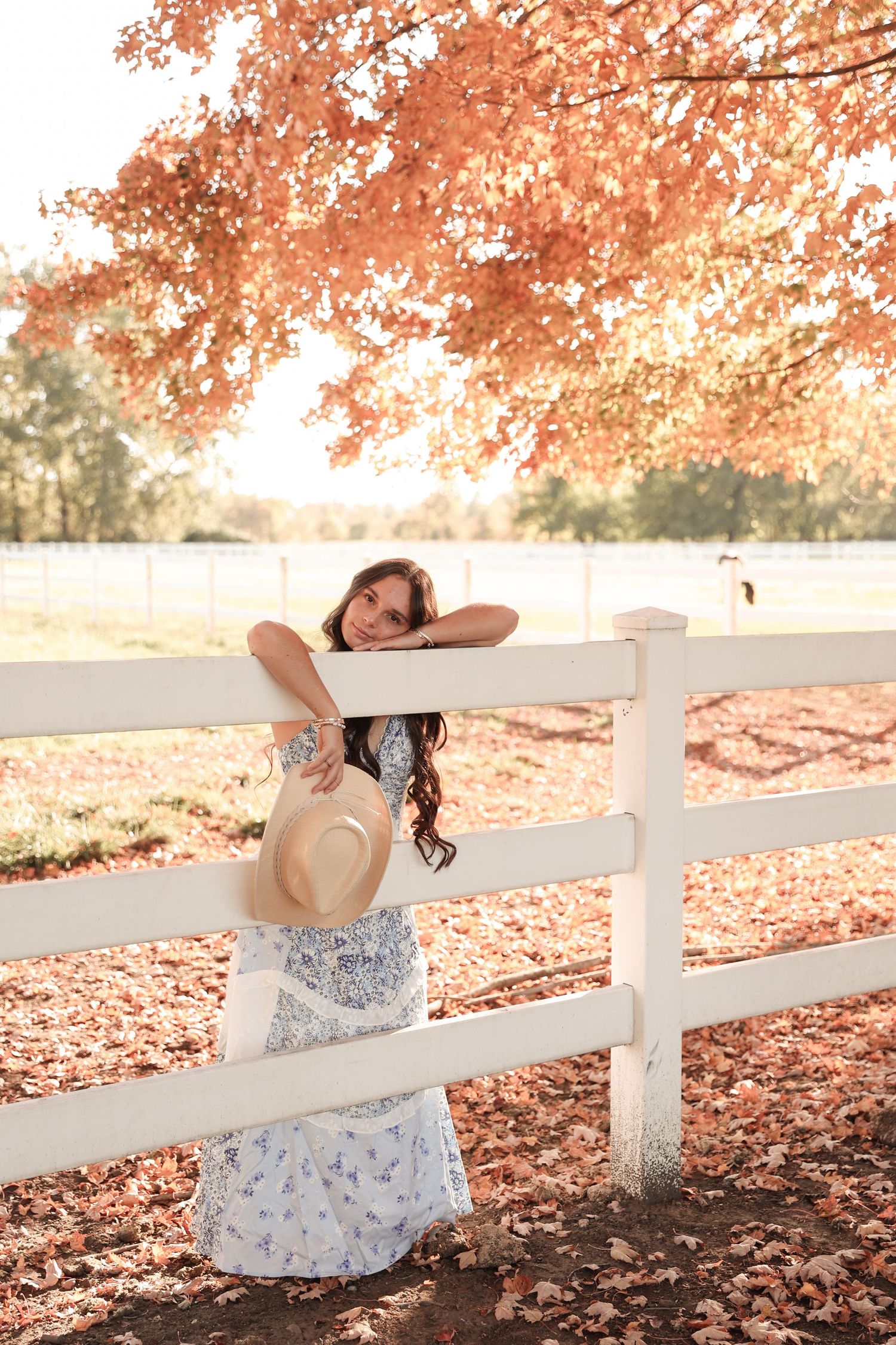 Woman in floral dress leaning on white fence under autumn red maple tree on a farm.