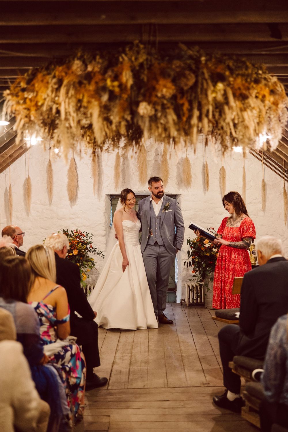 Romantic wedding ceremony with suspended floral decorations and guests seated in a rustic indoor venue.