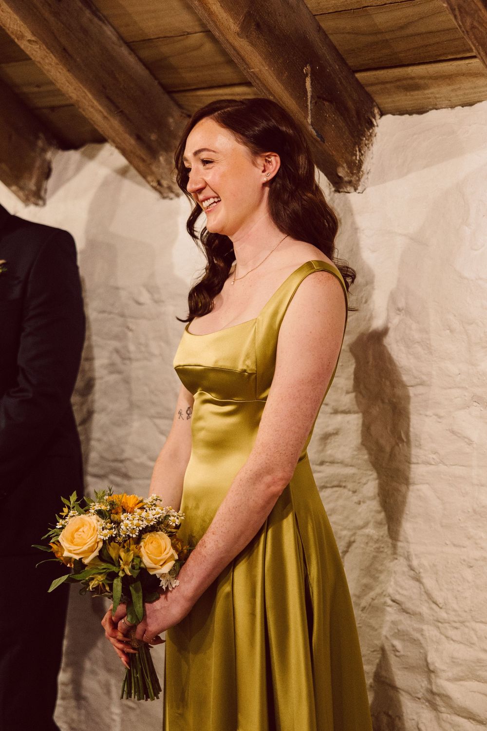 A person in a chartreuse satin bridesmaid dress smiles while holding a yellow rose bouquet against a white wall.