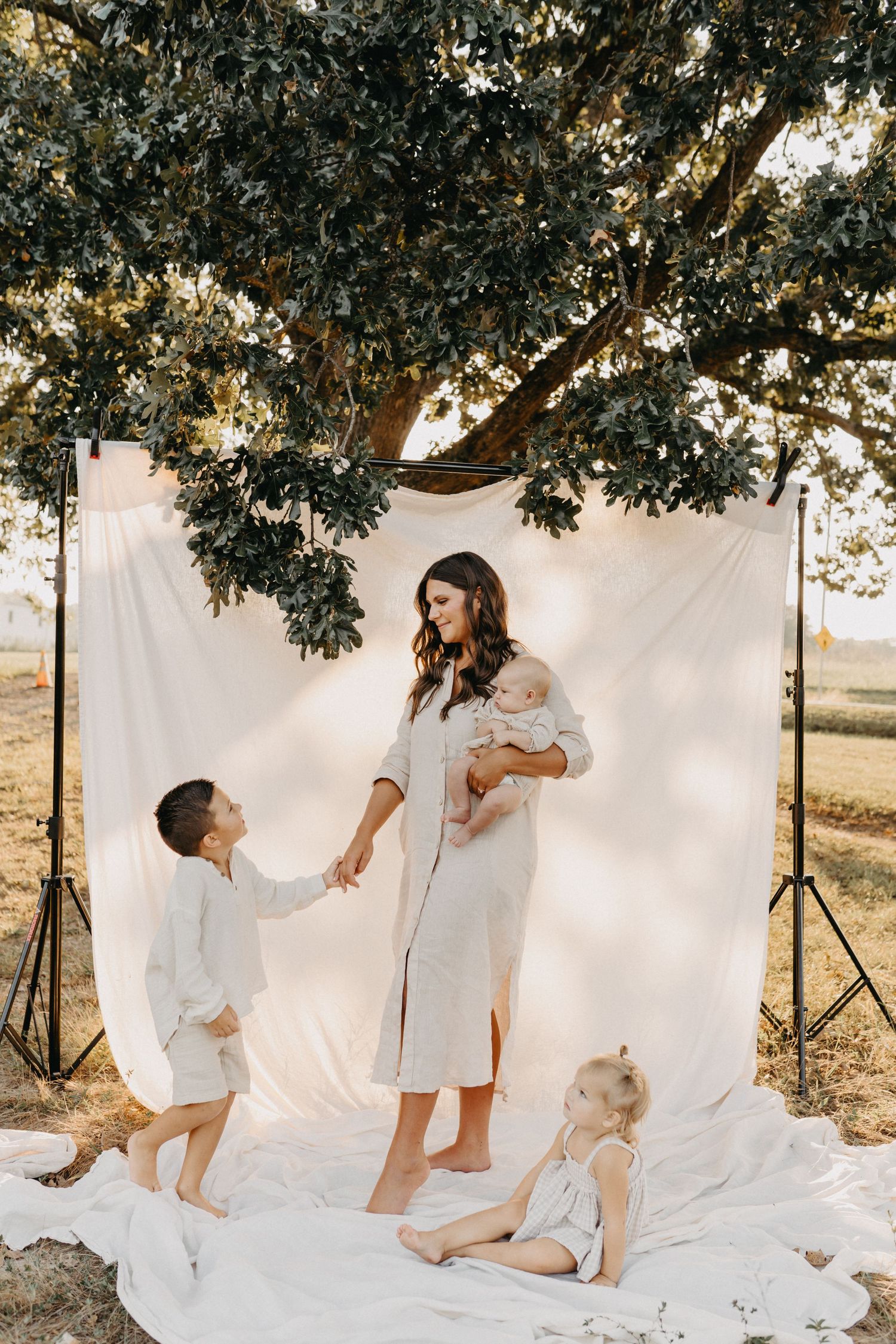 Mother holding baby outdoors with two children near white backdrop under large tree in natural light.