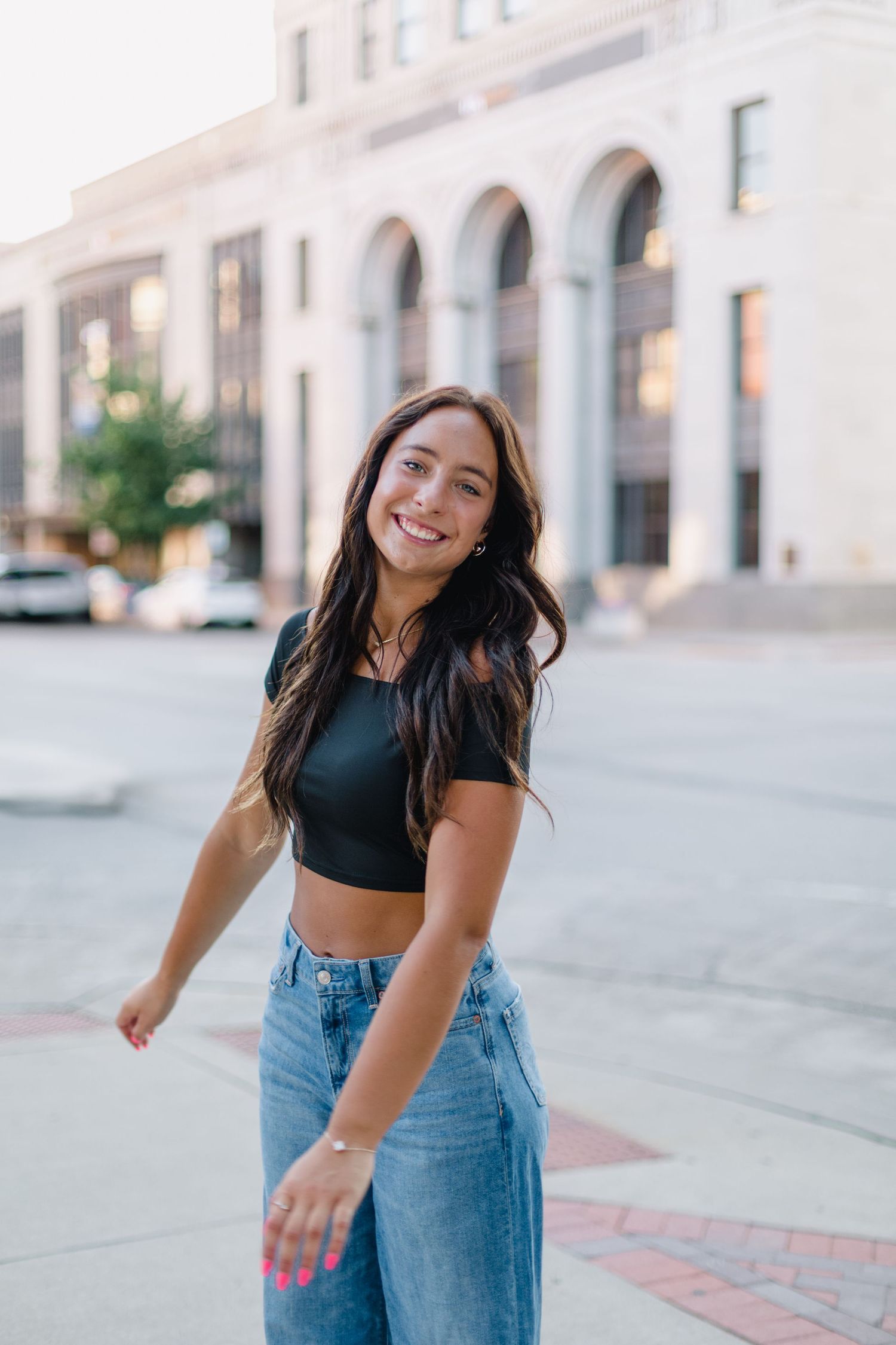 High school senior girl in black crop top and blue jeans poses playfully in downtown Cedar Rapids, IA with white buildings in background.